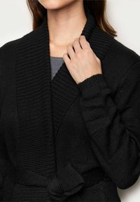 Czarny sweter dzianinowy z fakturowanym wzorem, szerokim kołnierzem szalowym i wiązanym paskiem w talii. Sweter ma długie rękawy.