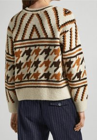 Persona che indossa un maglione crema con motivi geometrici marroni e arancioni e pantaloni navy a righe, vista da dietro.