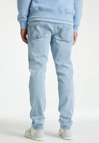 Svijetloplave traperice s taperiranim krojem, dizajn s pet džepova i prepoznatljivo šivanje stražnjih džepova, uparen s svijetlosivim cipelama.