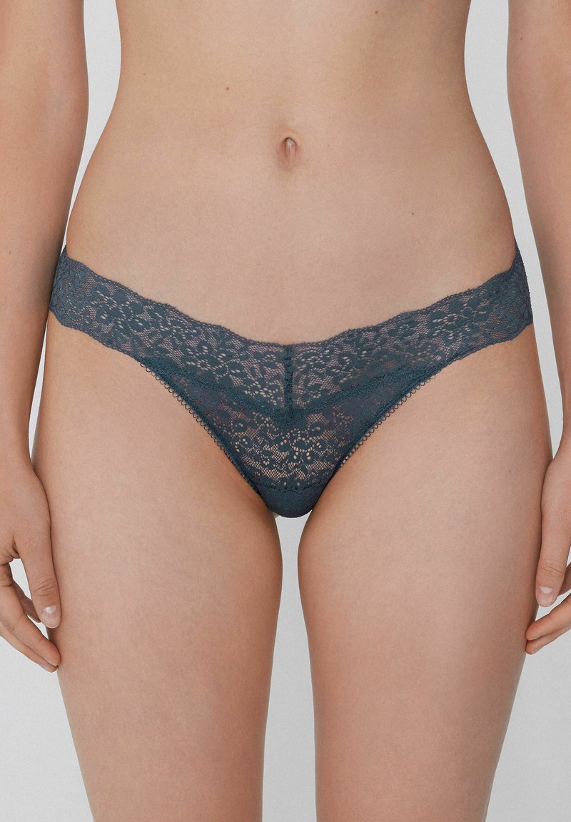 String en dentelle de teinte bleu sarcelle foncé, avec une ceinture festonnée et un motif floral. Texture douce avec des détails complexes sur tout le tissu.