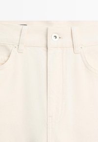 Primo piano della vita di jeans di colore crema con passanti per cintura, bottone in metallo e bordi superiori delle tasche anteriori.