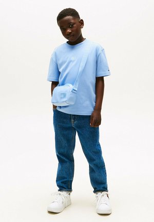 T-shirt bleu clair, sac bandoulière bleu, jean en denim bleu et baskets blanches. Le t-shirt présente un petit logo sur la poitrine.