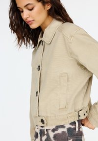 Giacca di jeans beige a vita corta con colletto, bottoni frontali e tasche laterali. Presenta dettagli di cucitura e lati regolabili.