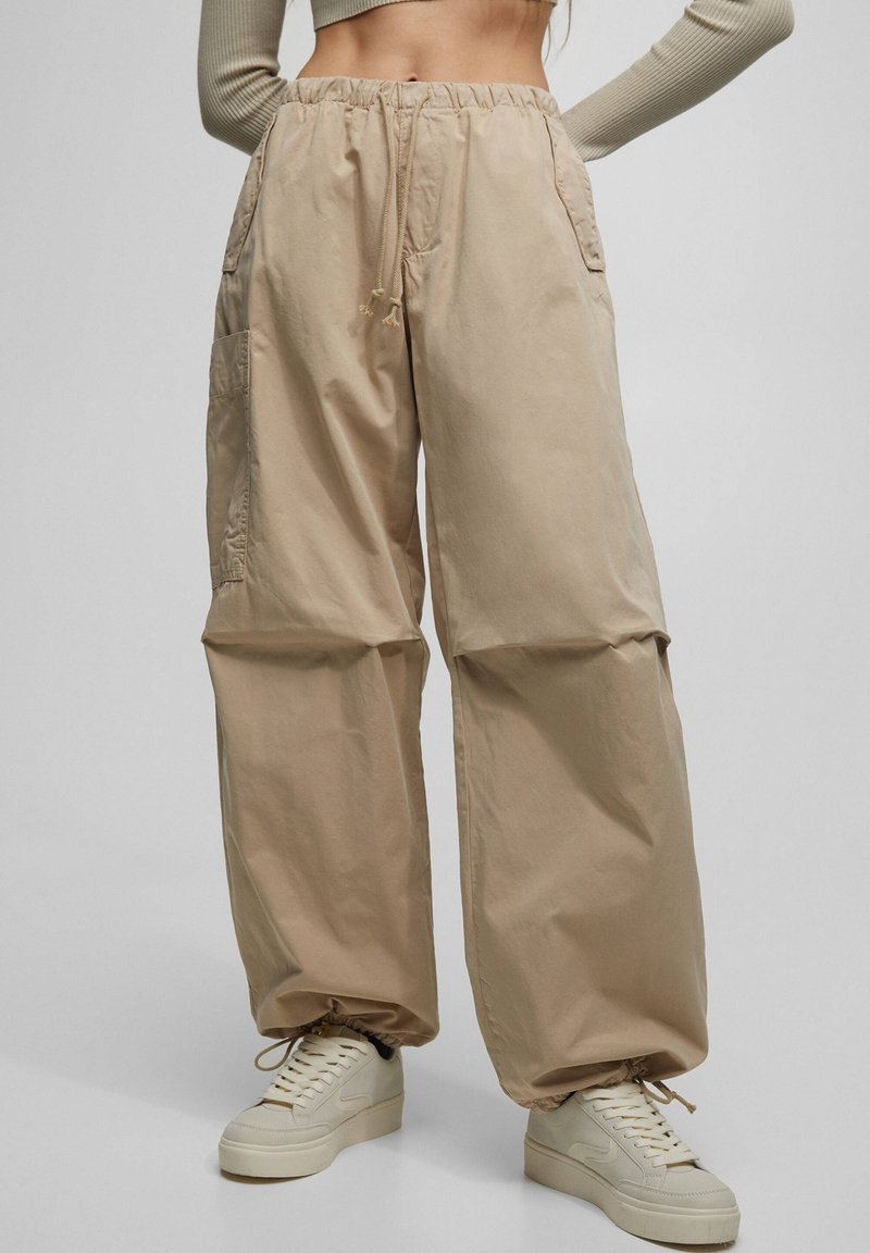 Pantalon cargo beige à jambes larges avec poignets et taille élastiques, assorti à des baskets crème sur un fond neutre.