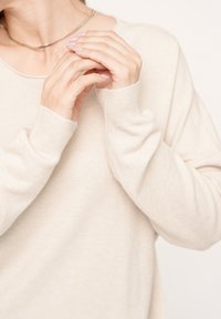 Beige Langarmpullover mit gerippten Bündchen, in entspannter Passform und mit weicher Textur. Hände zusammengelegt, mit hellrosa Nagellack.