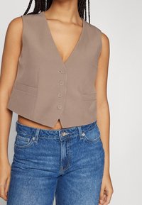 Brun ärmlös väst med djup V-hals, femknäppning och två sidofickor, i kombination med blå denimjeans. Slät textur.