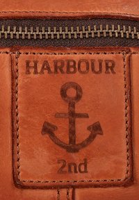 Pezzo in pelle marrone con testo in rilievo "HARBOUR" e un design centrale a ancora. Dettagli di cucitura evidenti lungo i bordi.