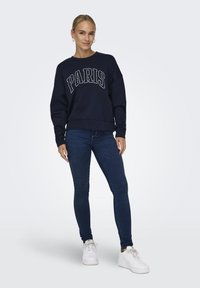 Tumši zils sporta krekls ar balto grafiku "PARĪZE", ribotās malas un brīvs piegriezums, apvienots ar tumši zilām šaurām biksēm un baltām kedām.