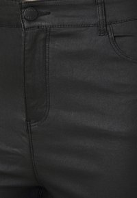 Gros plan d'un jean en denim noir montrant la ceinture, le bouton, les surpiqûres et le bord de la poche avant.