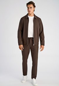 Bruine jas met ritssluiting en kraag, gecombineerd met bijpassende bruine joggers en witte sneakers; eenvoudig ontwerp, gladde textuur, zonder patronen.