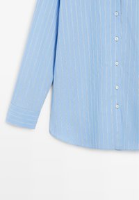 Blå langærmet skjorte med tynde hvide lodrette nålestriber og hvide knapper ned foran, manchetærme synlig på venstre side.