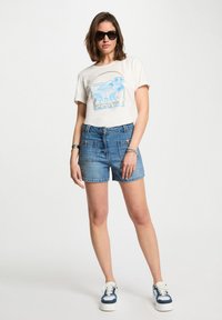 T-shirt bianca con grafica blu di sedie e un ombrello, abbinata a pantaloni corti di jeans blu. Sneakers bianche con dettagli blu completano il look.