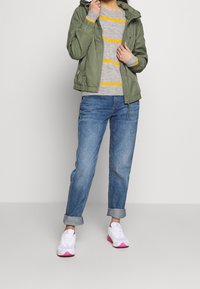 Olivengrøn lynlåstrøje med hætte, grå stribet sweater, opsmøgede denimjeans og hvide sneakers med pink detaljer.