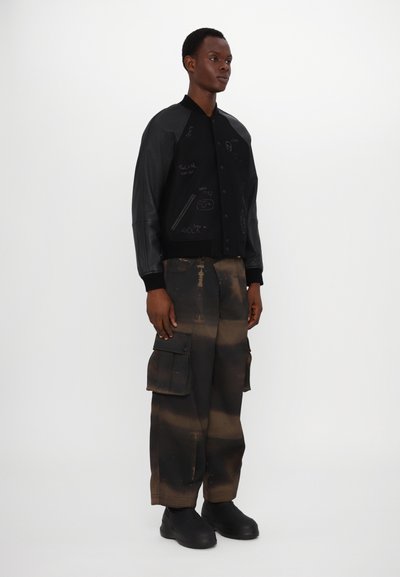 Veste bomber noire avec des manches en cuir, ornée de divers dessins manuscrits. Associée à un pantalon cargo marron et noir, et des bottes noires.