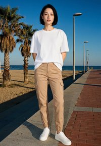 Junge Frau in weißem T-Shirt, beigen Cargohosen und weißen Turnschuhen steht auf einem Weg neben Palmen und dem Meer unter klarem blauem Himmel.