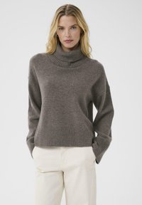 Brown knit turtleneck sweater with ribbed cuffs and hem. Soft texture, relaxed fit, and dropped shoulders. Paired with light beige pants.