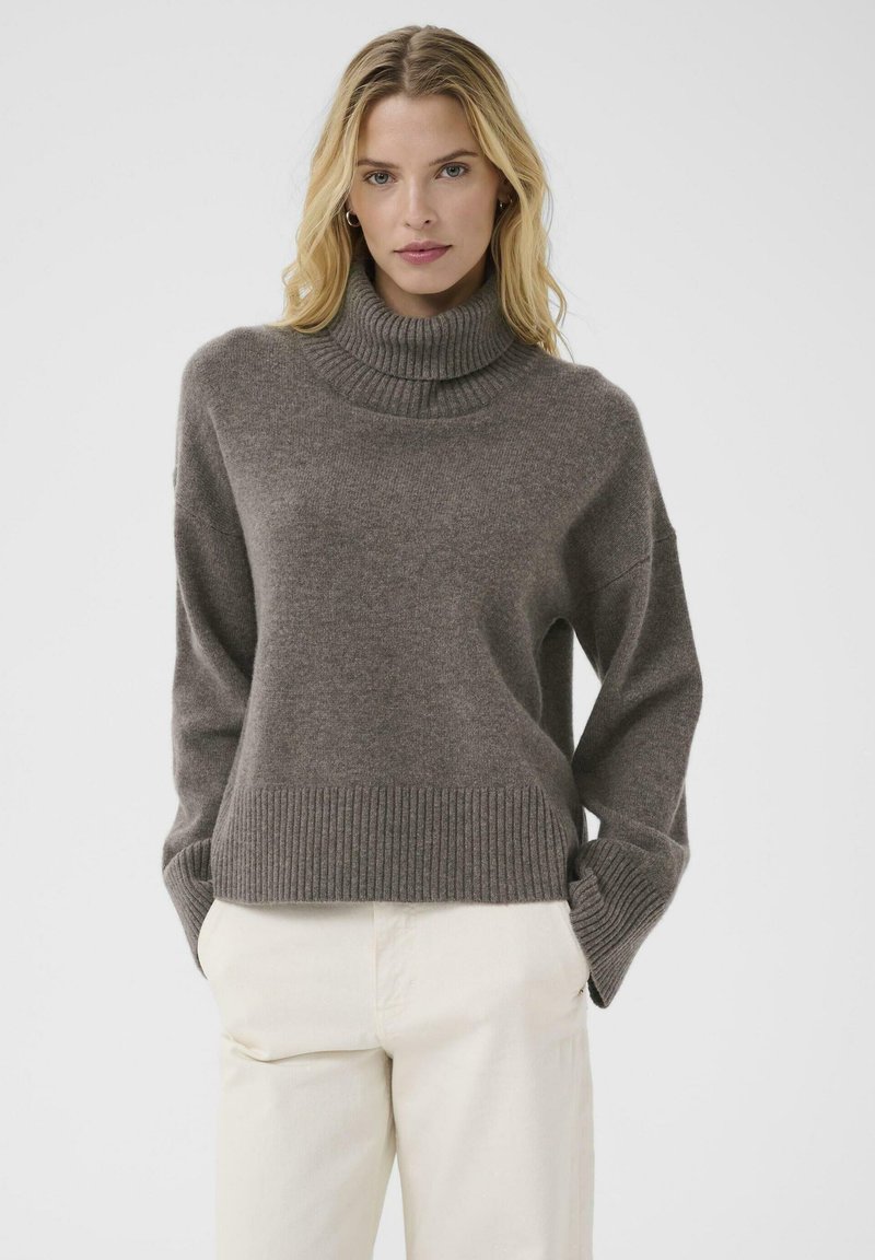 Brown knit turtleneck sweater with ribbed cuffs and hem. Soft texture, relaxed fit, and dropped shoulders. Paired with light beige pants.