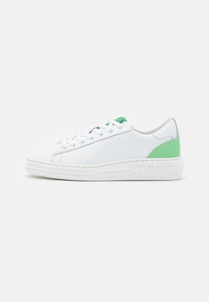Baskets basses blanches avec un patch vert au talon et un détail sur la languette, lacets blancs et semelle épaisse avec marquage en relief sur le côté.