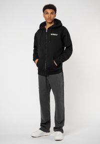 Black zip-up hoodie with front pockets and drawstring, paired with gray cargo pants. White sneaker accents visible. Simple, functional design.