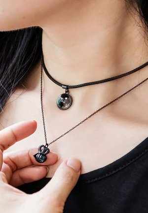 Femme portant deux colliers noirs, l'un avec un pendentif rond et l'autre avec un pendentif en forme d'abeille qu'elle tient dans sa main.
