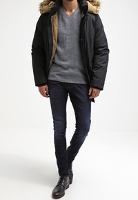 Giacca invernale nera con cappuccio foderato in pelliccia, maglione grigio, jeans blu e stivali in pelle marrone. Presenta una chiusura con zip e tessuto testurizzato.