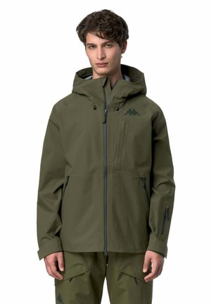 Giovane uomo con giacca con cappuccio verde oliva e pantaloni cargo abbinati, in piedi davanti a uno sfondo bianco.