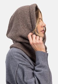 Kobieta w brązowym dzierganym kapturze i szarym swetrze, poprawiająca kaptur ręką, stojąca bokiem z zamkniętymi oczami.