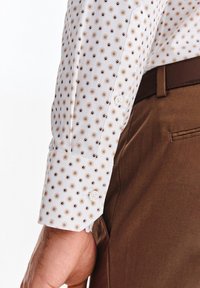 Biała koszula z małymi brązowymi i granatowymi wzorami, z dwoma guzikami przy mankietach, zestawiona z brązowymi spodniami. Tekstura wydaje się gładka.