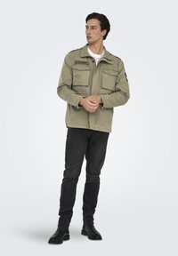 Giacca verde oliva in stile militare con due tasche frontali, spalline e la scritta "U.S. ARMY". Indossata sopra una maglietta bianca e jeans neri.