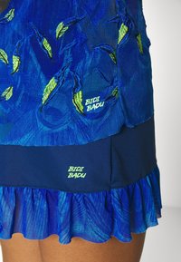 Modrá šaty s vyšívanými zelenými listovými vzory. Mají volánkový lem a neonové loga v dolní části. Vyrobeno z lehké, texturované tkaniny.