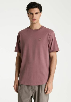 Jonge man staand, met een mauve korte mouw t-shirt en een grijze broek tegen een effen witte achtergrond.