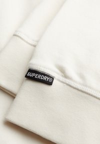 Tela de sudadera blanca con puños acanalados texturizados. Presenta una etiqueta negra "SUPERDRY" cosida en el puño. Diseño suave y casual.
