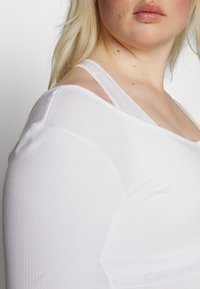 T-shirt branca de manga longa com um design à mostra os ombros, apresentando tiras finas e uma textura suave.