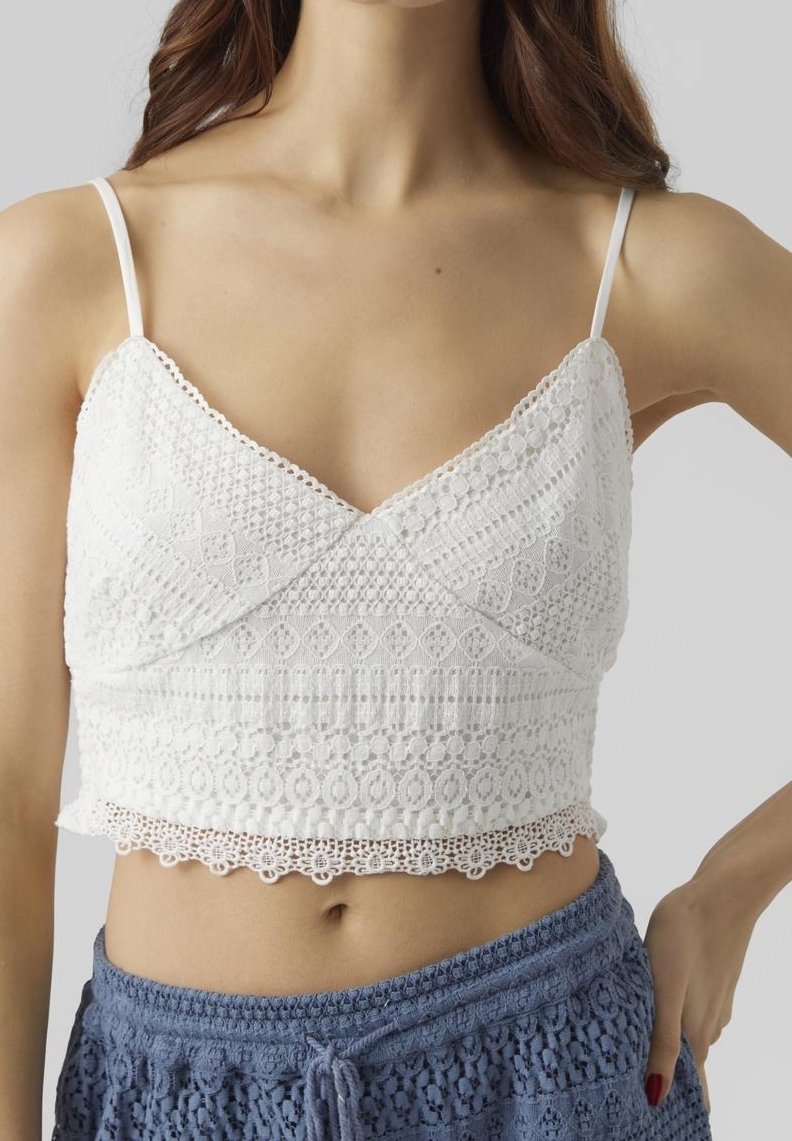 Femme portant un crop top en dentelle blanche à fines bretelles et un short bleu au crochet, debout, une main sur la hanche, sur fond uni.