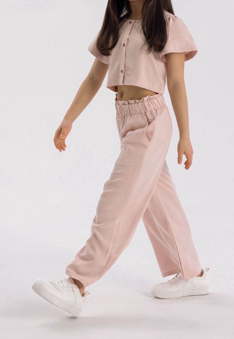 Jeune personne marchant, portant un top court rose clair boutonné, un pantalon taille haute assorti et des baskets blanches, montrée en plein pas sur fond uni.