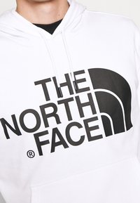Felpa bianca con cappuccio e lacci neri, caratterizzata da un grande testo nero "THE NORTH FACE" e dal logo a mezzo cupola del marchio sul petto.