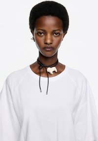 Collar negro con un charm decorativo blanco, llevado sobre un sudadera blanca amplia. El charm se asemeja a una figura animal abstracta.