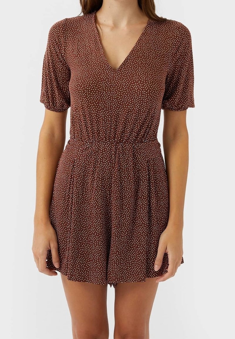 Femme portant une combinaison courte marron à manches courtes avec des pois blancs, un col en V et une taille élastique.