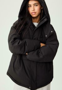 Mlada žena s dugom tamnom kosom nosi crnu oversized puffer jaknu s kapuljačom, s prekriženim rukama na neutralnoj pozadini.