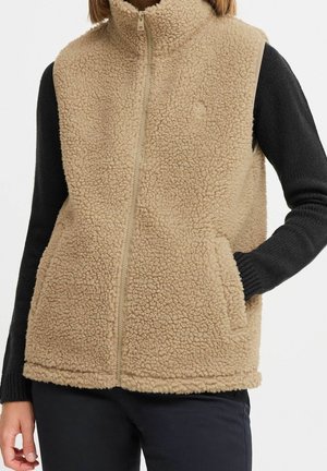 Beige sherpa-fleecevest med lynlås, båret over en sort langærmet trøje med hænderne i vestens lommer.