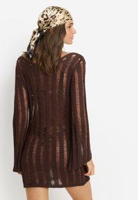 Καφέ πλεκτό φόρεμα με κάθετες ρίγες και φαρδιά γραμμή. Το μοντέλο φοράει μεταξωτό μαντήλι με λεοπάρ σχέδιο δέμενο στο πίσω μέρος του κεφαλιού της.