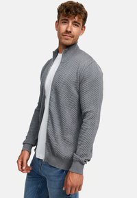 Grå zip-up cardigan med en teksturert herringbone-mønster, ribbestrikkede mansjetter og kant, brukt over en hvit t-skjorte og kombinert med blå jeans.