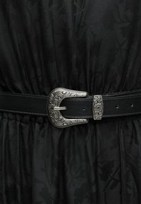 Μαύρη δερμάτινη ζώνη με λεπτομερή ασημένια αγκράφα με λουλουδένια χάραξη και θηλιά, φορεμένη πάνω από ρούχο από μαύρο υφασμάτινο ύφασμα με υφή.