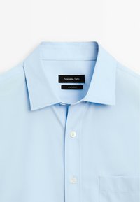 Camicia azzurra chiara con colletto classico, apertura a bottoni frontale, taschino singolo e etichette che mostrano il nome del marchio Massimo Dutti, prodotta in Egitto.