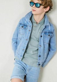 Ljusblå denimjacka med framfickor, kombinerad med en dämpad grön polo och ljusblå denimshorts. Svarta solglasögon fulländar looken.