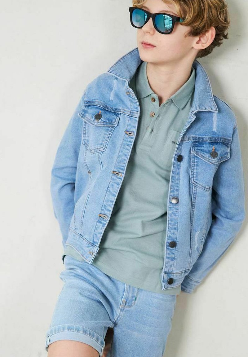 Ljusblå denimjacka med framfickor, kombinerad med en dämpad grön polo och ljusblå denimshorts. Svarta solglasögon fulländar looken.