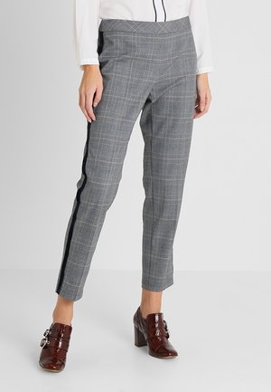 Femme portant un pantalon slim à carreaux gris avec des bandes latérales noires et des mocassins à talons marron, debout devant un fond uni.