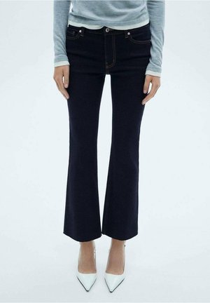 Persoon met zwarte uitlopende broek, lichtgrijs langemouwen shirt en witte spitse hoge hakken, staand tegen een effen achtergrond.