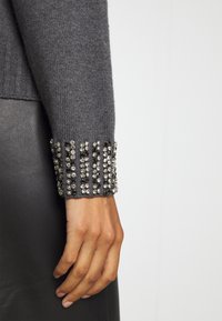 Main à la peau foncée portant une manche tricotée grise ornée de rangées verticales de strass clairs sur le poignet.