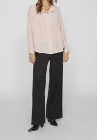 Blusa rosa chiaro con scollo a V e davanti con bottoni, abbinata a pantaloni neri a gamba larga. La blusa presenta maniche lunghe e una texture leggera.